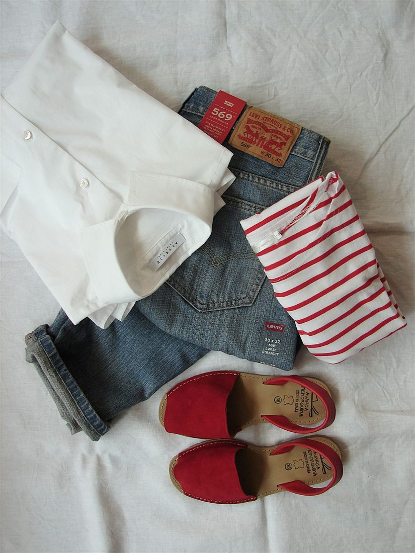 A flat lay on a pale sheet: a white shirt, faded blue jeans, a red and white striped top and a pair of red sandals.