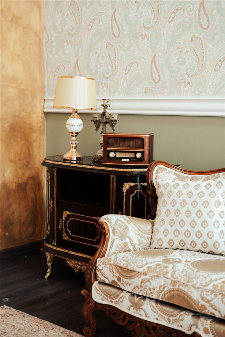 A sitting room: pale paisley wallpaper above an olive lower wall, a golden wall on the left, a dark gilded cabinet with a lamp and an old radio, and a cream sofa with a carved wooden frame.